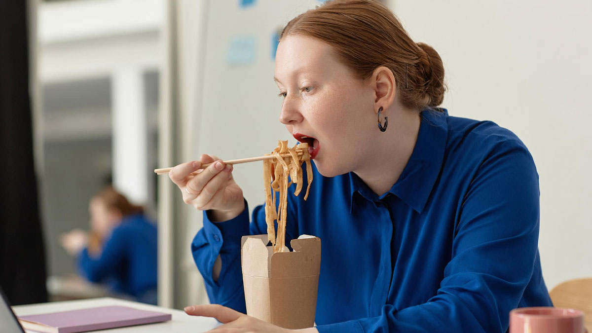 Woman eating noodles at office desk, illustrating workers adding labels to protect their food from lunch thief.
