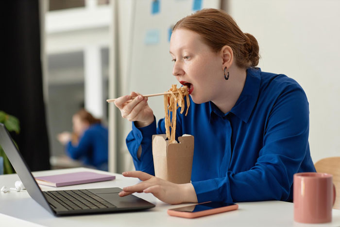Woman eating takeout noodles while working on laptop, illustrating workers adding labels to protect their food from lunch thieves. Woman eating takeout noodles while working on laptop, illustrating workers adding labels to protect their food from lunch thieves.