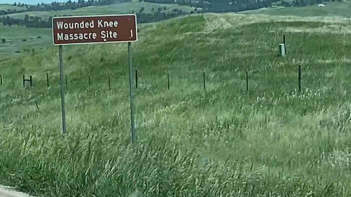 Roadside sign for Wounded Knee M******e Site surrounded by green fields, a creepy and chilling spot in the U.S.