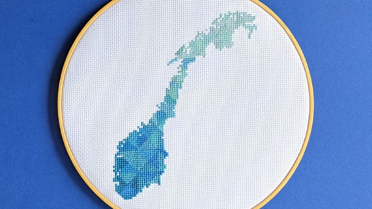 Cross-stitched map of Norway in shades of blue and green inside an embroidery hoop on a blue background.