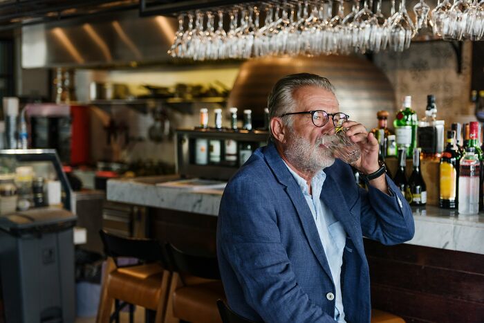 Older man in glasses sitting at bar counter drinking water among bottles, relating to dates that went sour and safe word use.