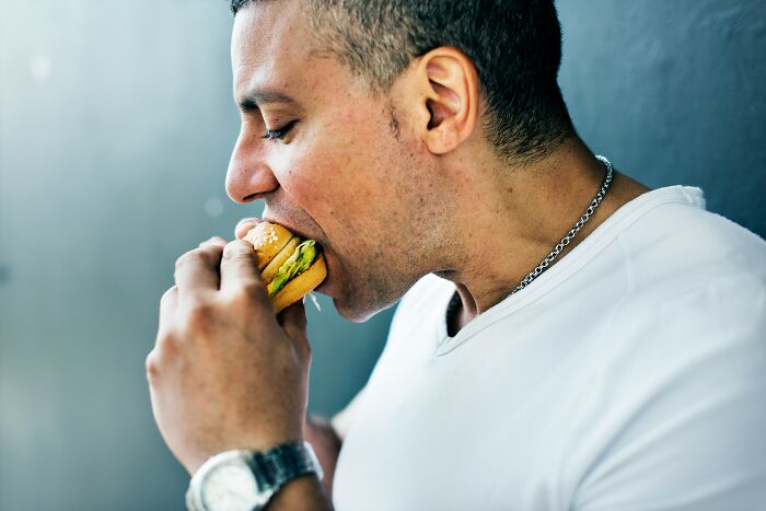 Man in white shirt eating a sandwich while thinking about coworker diabolical things to report to HR.