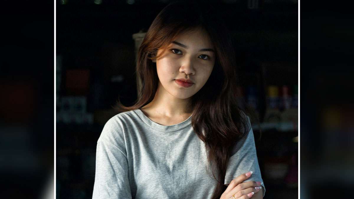 Young woman with long hair and neutral expression, representing raw truths about human nature in a dimly lit setting.