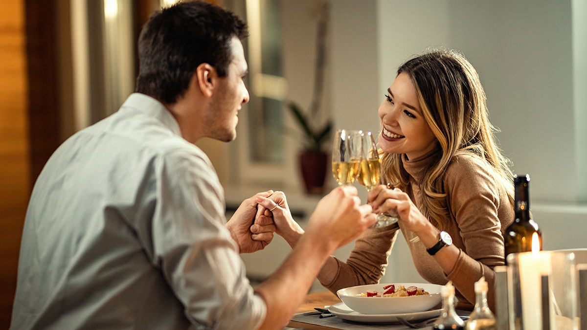 Couple on a first date holding hands and toasting wine glasses over dinner in a cozy restaurant setting.
