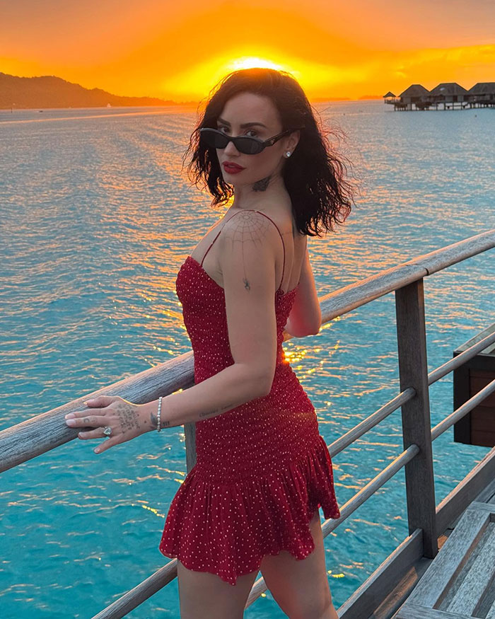 Demi Lovato posing in a red dress by the ocean at sunset, with sunglasses and tattoos visible on her arm. Demi Lovato posing in a red dress by the ocean at sunset, with sunglasses and tattoos visible on her arm.