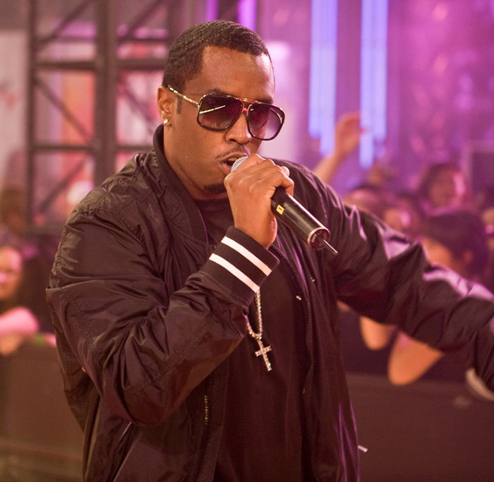 Diddy performing on stage wearing sunglasses and a black jacket, highlighting his aged look during a court trial.