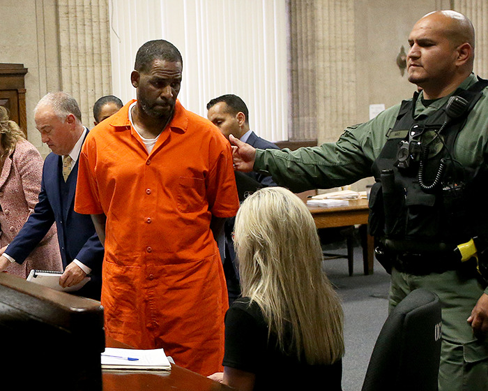 Rapper R Kelly in orange prison uniform in courtroom with guard and legal team during trial proceedings. Rapper R Kelly in orange prison uniform in courtroom with guard and legal team during trial proceedings.