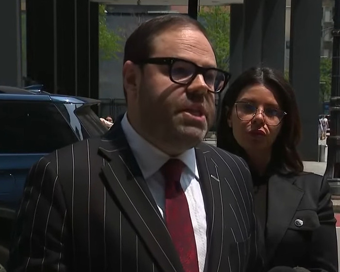 Man in pinstripe suit and red tie speaking outdoors with woman in black jacket and glasses behind him. Man in pinstripe suit and red tie speaking outdoors with woman in black jacket and glasses behind him.