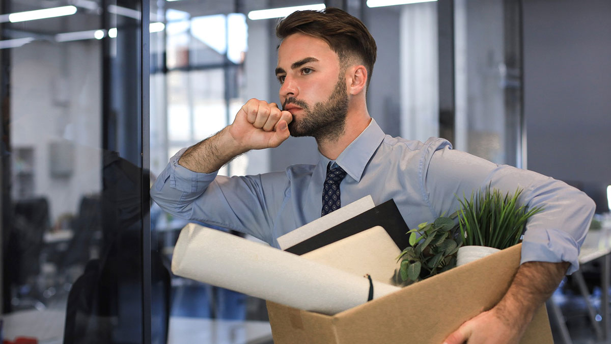 Young worker holding a box of office belongings, looking thoughtful and concerned after losing his job in a modern office.