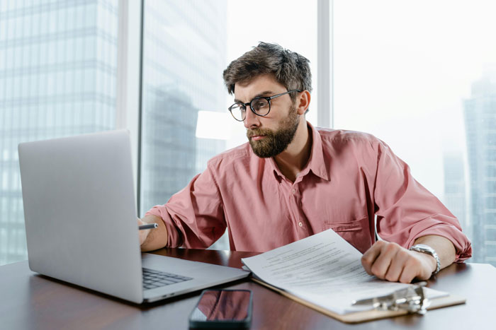Man in glasses working on laptop at office desk with documents, reflecting on career advice and job challenges.