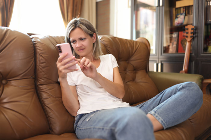 Stepmom sitting on couch looking upset and frustrated while using a smartphone in a cozy living room setting Stepmom sitting on couch looking upset and frustrated while using a smartphone in a cozy living room setting