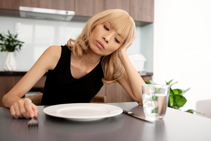 Young woman looking tired and weak while sitting at a table with an empty plate, illustrating prolonged fasting dangers. Young woman looking tired and weak while sitting at a table with an empty plate, illustrating prolonged fasting dangers.