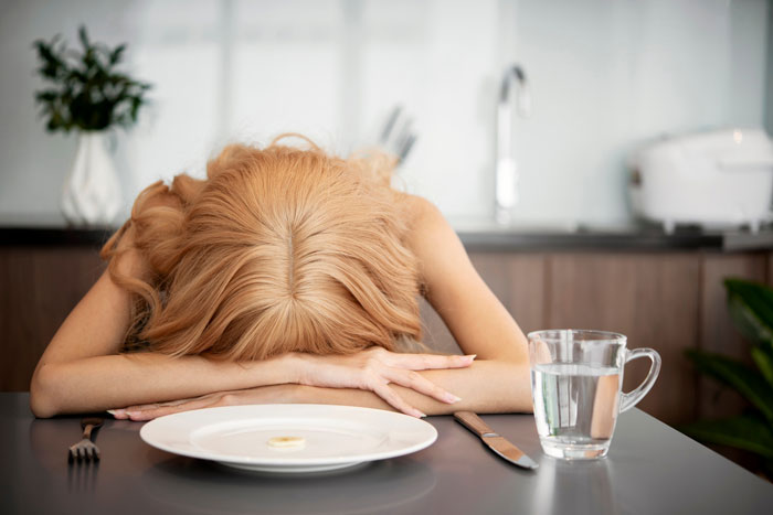 Woman resting head on table with a small piece of food on plate and glass of water, illustrating prolonged fasting risks. Woman resting head on table with a small piece of food on plate and glass of water, illustrating prolonged fasting risks.