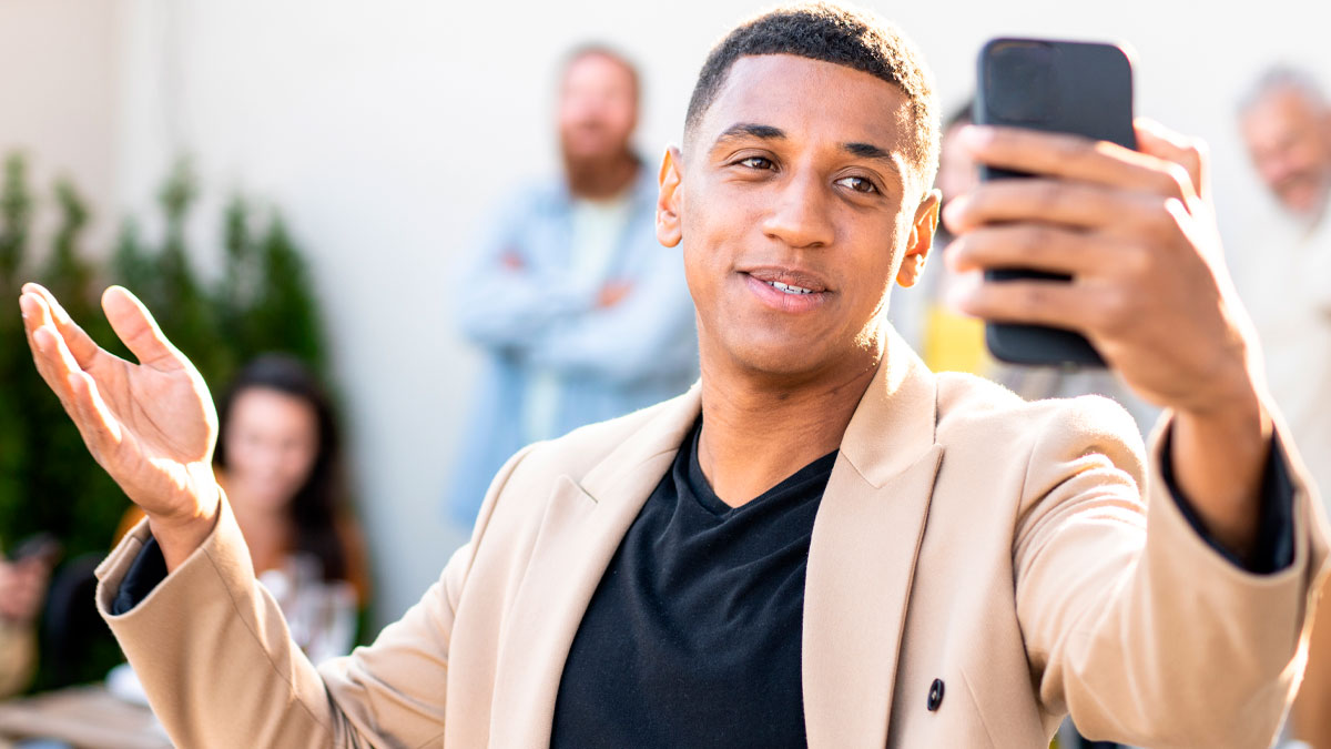 Young influencer marketing coworker taking a selfie, wearing a beige jacket, with coworkers blurred in the background.