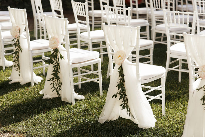 White wedding chairs decorated with flowers and greenery set up outdoors for a ring bearer and guest seating arrangement. White wedding chairs decorated with flowers and greenery set up outdoors for a ring bearer and guest seating arrangement.