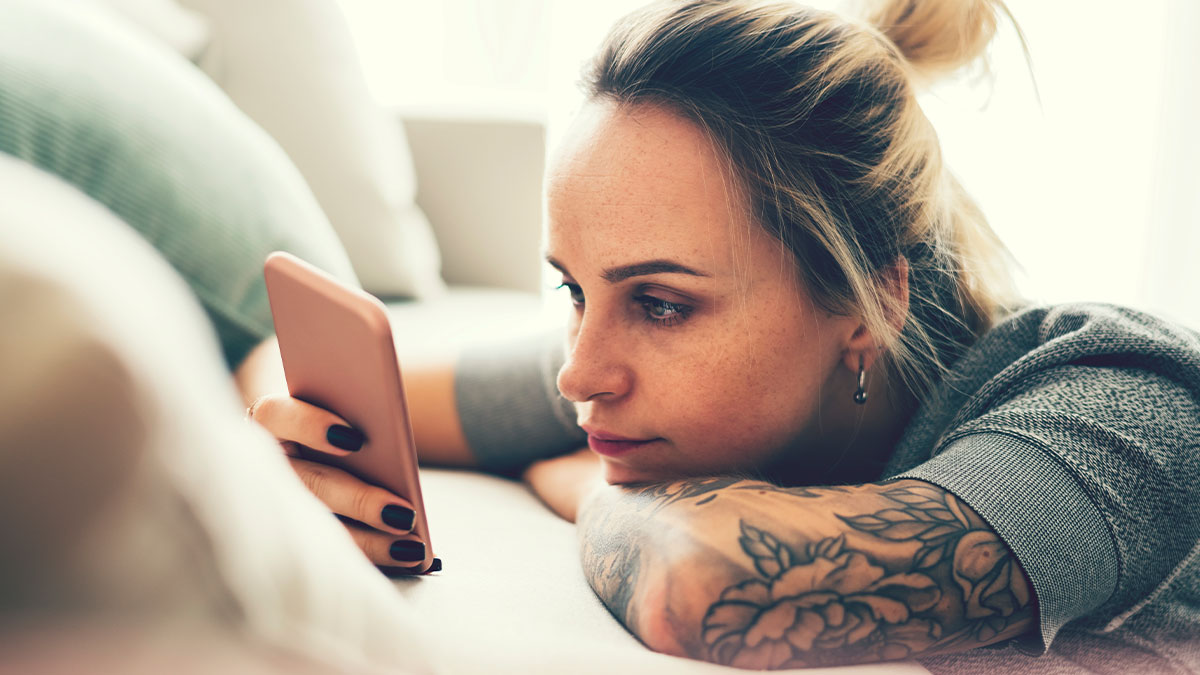 Young woman with tattooed arm lying on couch, looking at phone, reflecting on family RSVP no to wedding conflict.