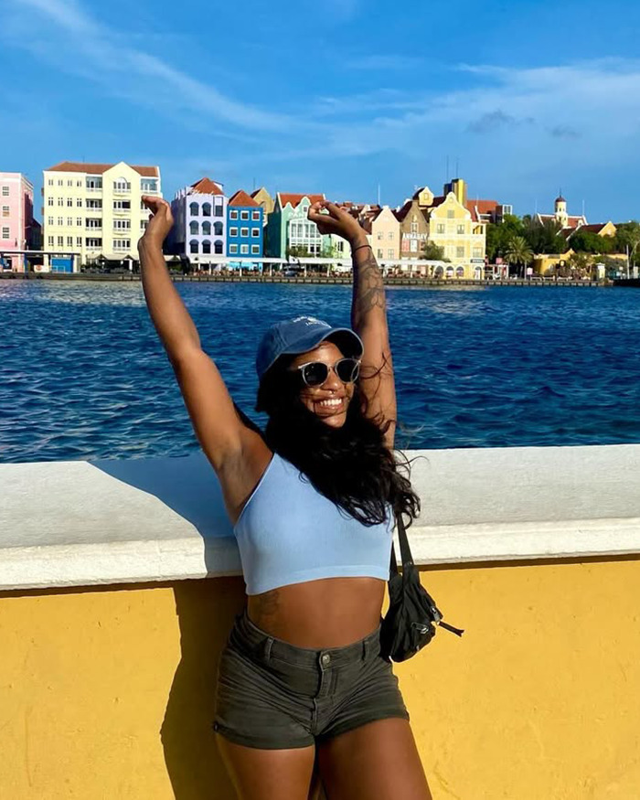Woman smiling with arms raised by waterfront, colorful buildings behind, related to tourist trapped inside active volcano. Woman smiling with arms raised by waterfront, colorful buildings behind, related to tourist trapped inside active volcano.