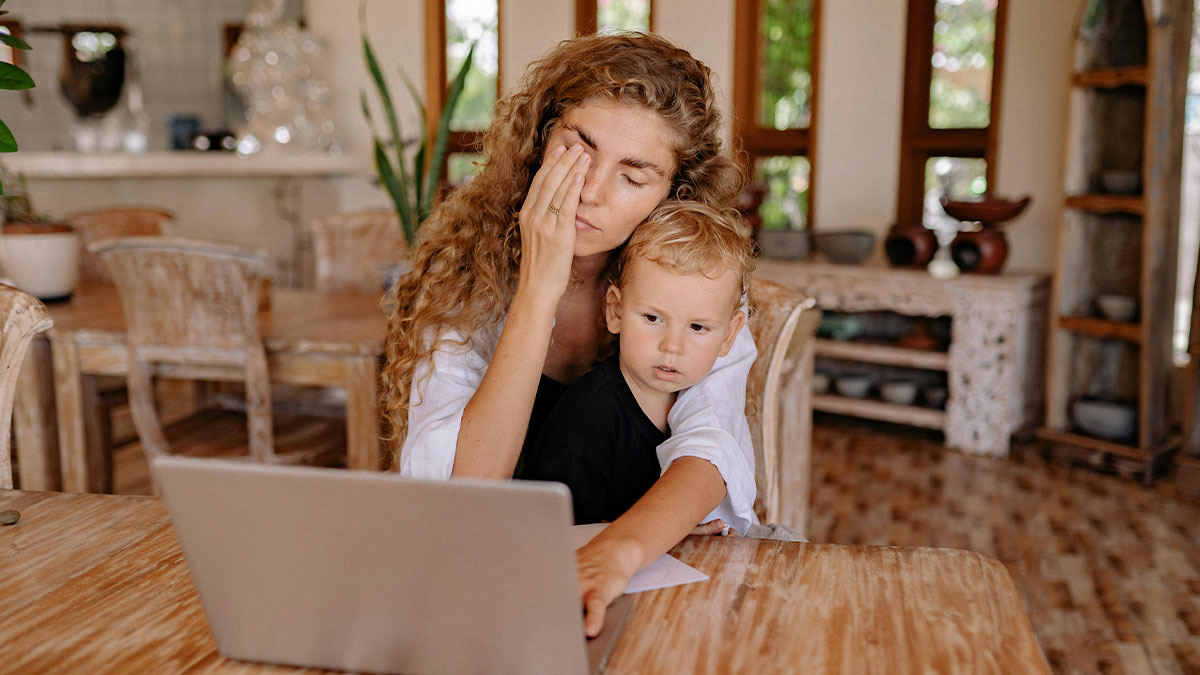 Woman juggling baby and online business looking stressed while working on laptop at home dining table.