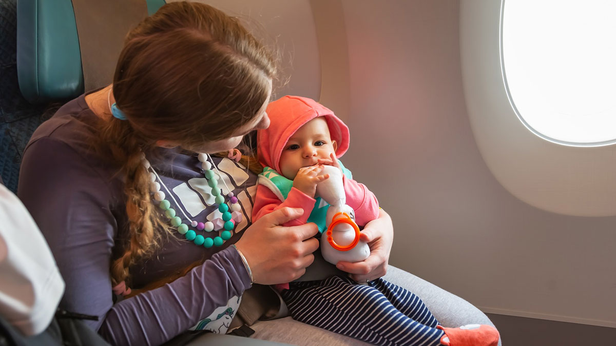 Mom nursing baby on a flight, holding toy, seated next to airplane window during in-flight service.