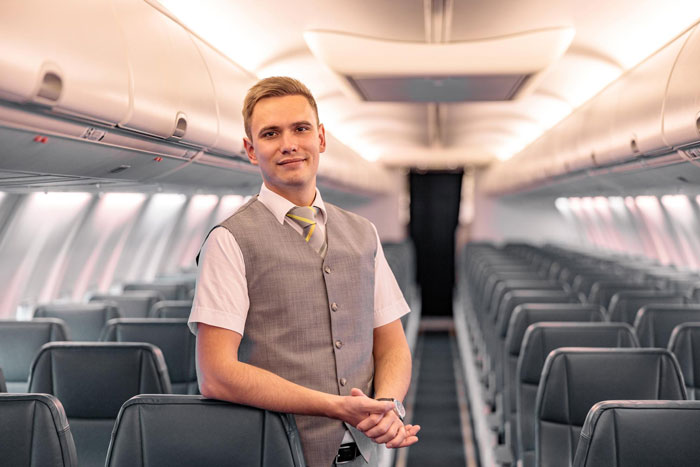Male steward standing in empty airplane cabin, wearing uniform vest and tie, ready to serve passengers during flight. Male steward standing in empty airplane cabin, wearing uniform vest and tie, ready to serve passengers during flight.