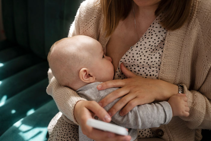 Mother breastfeeding baby while holding a phone, highlighting nursing cover and breastfeeding in a home setting. Mother breastfeeding baby while holding a phone, highlighting nursing cover and breastfeeding in a home setting.