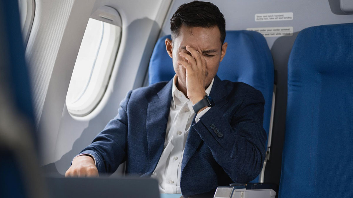 Man in business attire rubbing eyes feeling stressed on airplane, highlighting flight attendant safety questions.