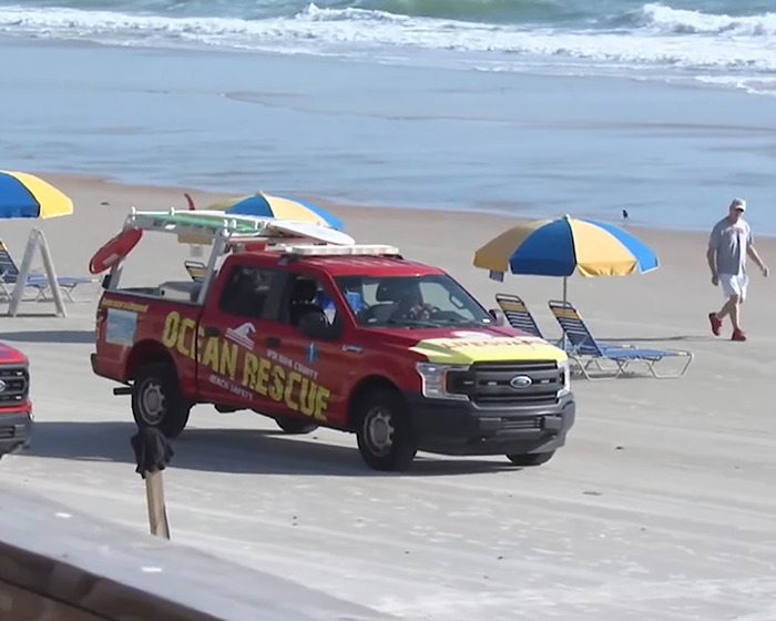 Red ocean rescue truck parked on a beach near umbrellas and lounge chairs by the ocean shore. Red ocean rescue truck parked on a beach near umbrellas and lounge chairs by the ocean shore.