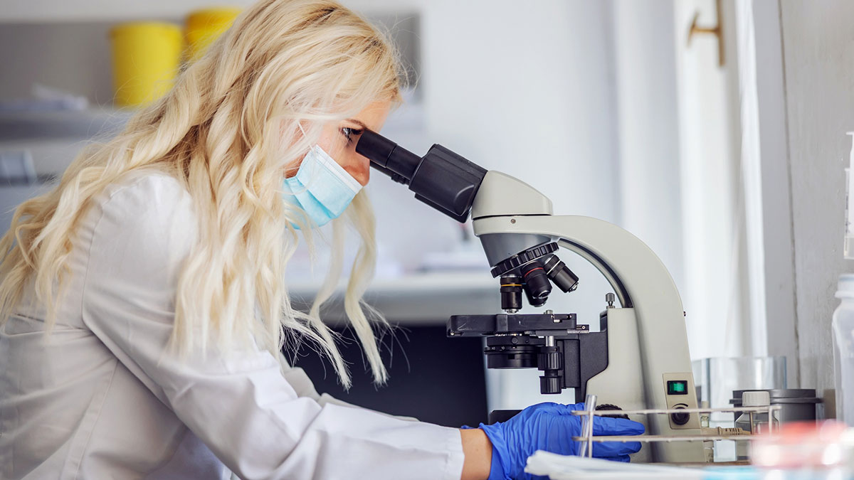 Scientist wearing a mask and gloves examining samples under a microscope studying fiber's effect on cancer-causing chemicals.