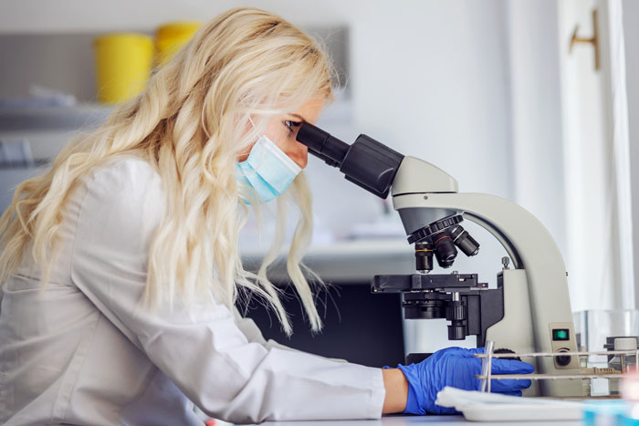 Scientist wearing mask and gloves examining samples under microscope in lab studying fiber effects on chemicals. Scientist wearing mask and gloves examining samples under microscope in lab studying fiber effects on chemicals.