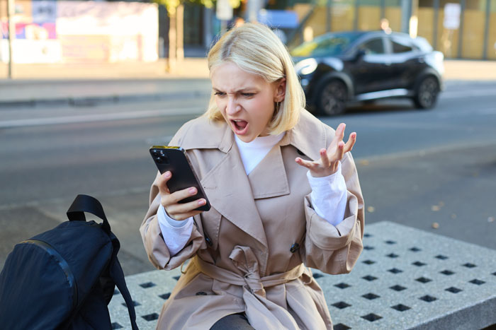 Angry woman sitting outdoors, frustrated while looking at phone, illustrating woman wants half of bestie's inheritance conflict.