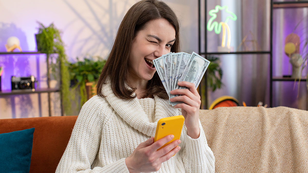 Woman holding cash and phone at home, representing woman wanting half of bestieu2019s inheritance after childhood struggles.