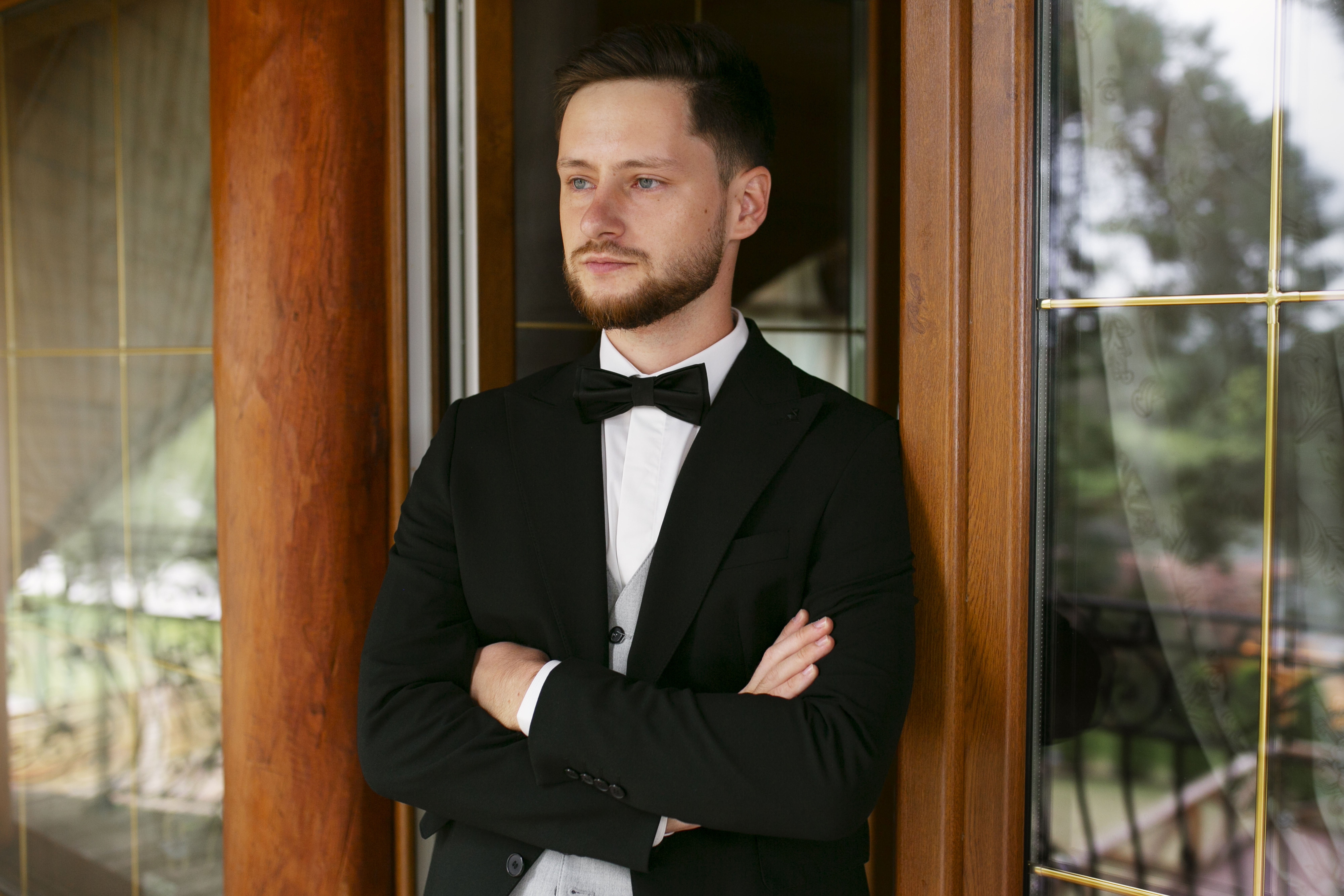 Best man in formal black suit and bow tie standing with arms crossed by a wooden door in wedding setting. Best man in formal black suit and bow tie standing with arms crossed by a wooden door in wedding setting.