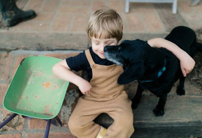 Young child sitting outdoors with a black dog, sharing a loving moment after a priceless reward from a wife.
