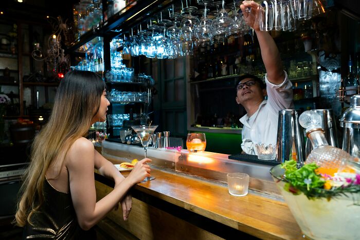 Woman sitting at bar holding cocktail glass while bartender reaches for hanging glasses in dimly lit setting, dates gone sour concept.