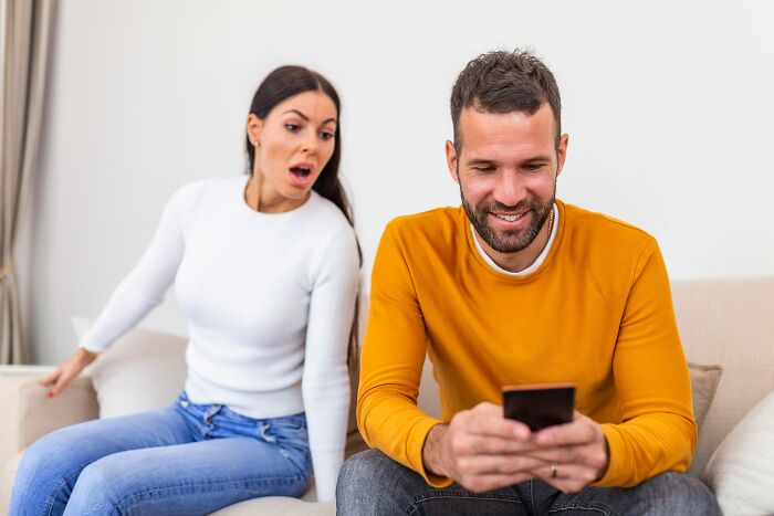 A woman showing surprise while a man smiles looking at his phone, illustrating signs of low self-esteem.