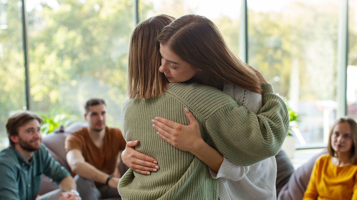 Two women hugging closely in a cozy room while others watch, showing a good trend of support slowly disappeared.