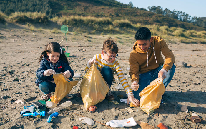 Three children cleaning a beach, collecting trash in yellow bags, showing a good trend slowly disappeared in environmental care.