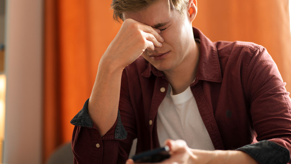 Young man stressed at workplace, holding phone and rubbing eyes, highlighting high school drama and hottest guy list issues.