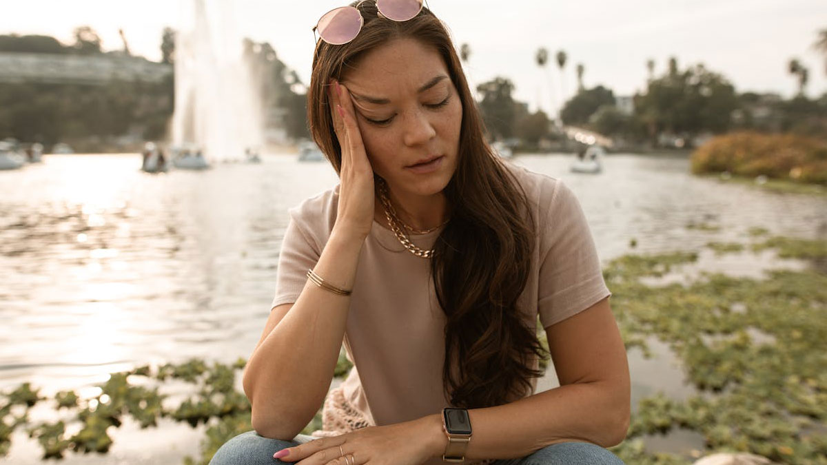 Young woman sitting by a lake looking upset, possibly reflecting on ruining boyfriend's perfect proposal plan due to being hangry.