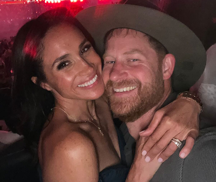Meghan Markle and Prince Harry smiling and embracing at a nighttime event with warm lighting and casual attire. Meghan Markle and Prince Harry smiling and embracing at a nighttime event with warm lighting and casual attire.