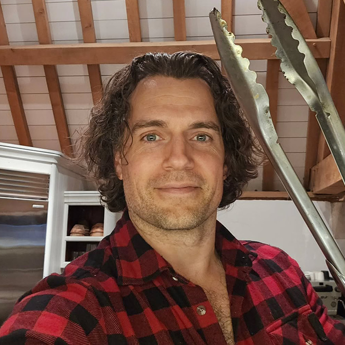 Henry Cavill with a new Highlander hairstyle, wearing a red plaid shirt and holding metal tongs indoors. Henry Cavill with a new Highlander hairstyle, wearing a red plaid shirt and holding metal tongs indoors.