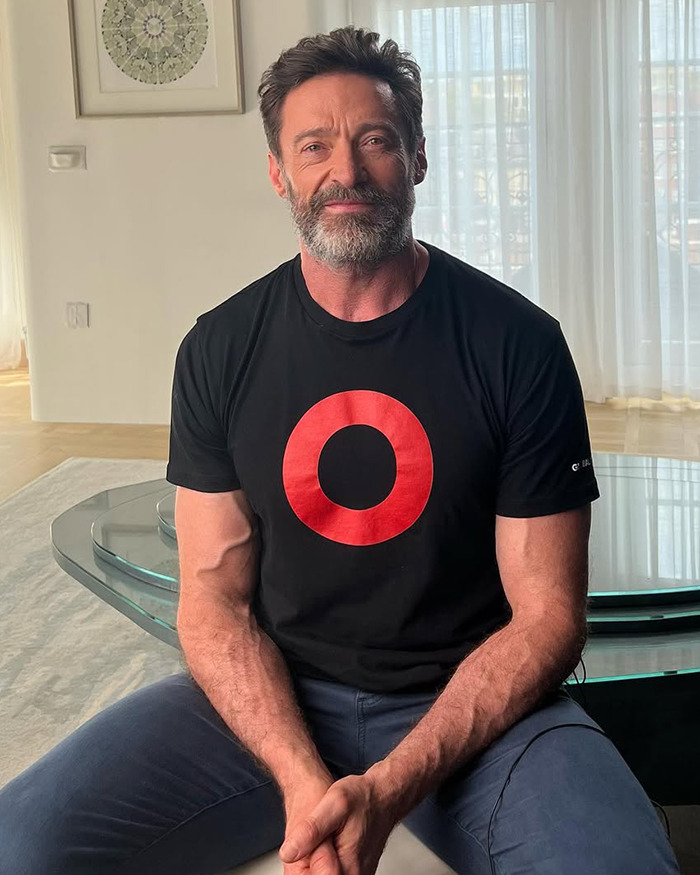 Hugh Jackman sitting indoors in casual black t-shirt with red circle, amid fans discovering 17-year history with Sutton Foster. Hugh Jackman sitting indoors in casual black t-shirt with red circle, amid fans discovering 17-year history with Sutton Foster.