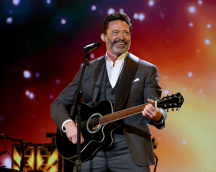Hugh Jackman performing on stage with guitar, smiling and dressed in a gray suit during a live show. Hugh Jackman performing on stage with guitar, smiling and dressed in a gray suit during a live show.