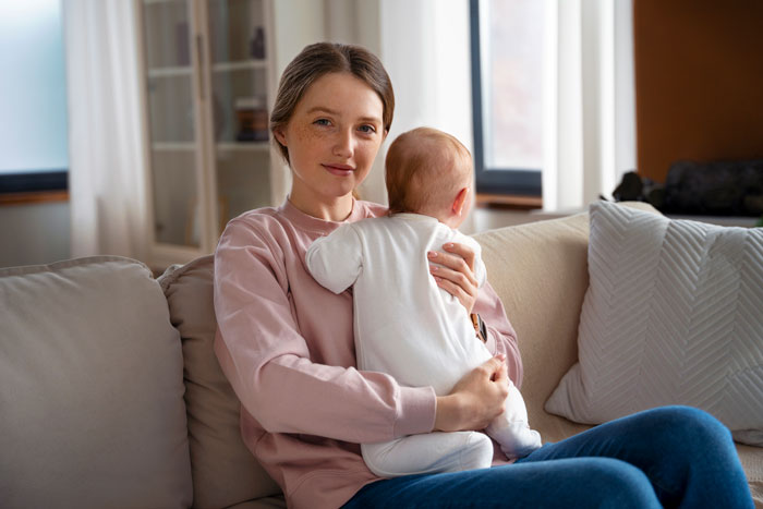 Mother holding baby on couch, highlighting wife sacrifice and one-sided effort in Sunday deal with husband enjoying lazy mornings. Mother holding baby on couch, highlighting wife sacrifice and one-sided effort in Sunday deal with husband enjoying lazy mornings.