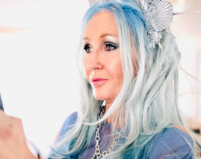 Woman with long blue hair and decorative headpiece, looking thoughtfully to the side in a bright indoor setting. Woman with long blue hair and decorative headpiece, looking thoughtfully to the side in a bright indoor setting.