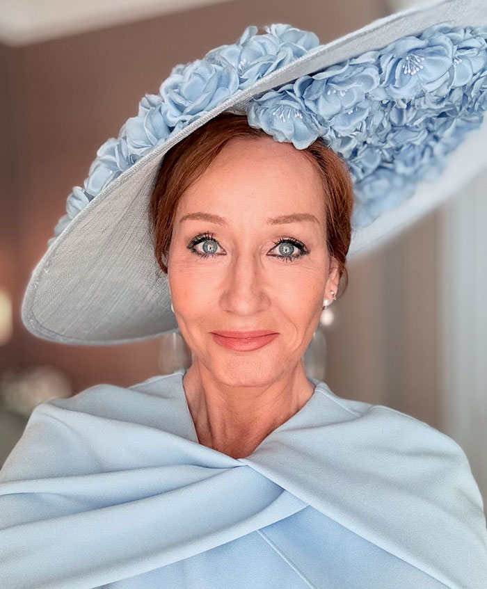 J.K. Rowling wearing a large blue floral hat and matching outfit, smiling softly in a well-lit room. J.K. Rowling wearing a large blue floral hat and matching outfit, smiling softly in a well-lit room.