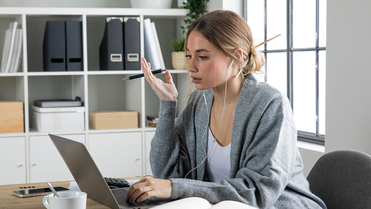 Young woman working on a laptop with earphones on, surrounded by office supplies in a modern workspace.