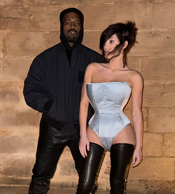 Bianca Censori wearing a daring bodysuit with hair extensions, standing next to a man in black clothing against a stone wall. Bianca Censori wearing a daring bodysuit with hair extensions, standing next to a man in black clothing against a stone wall.