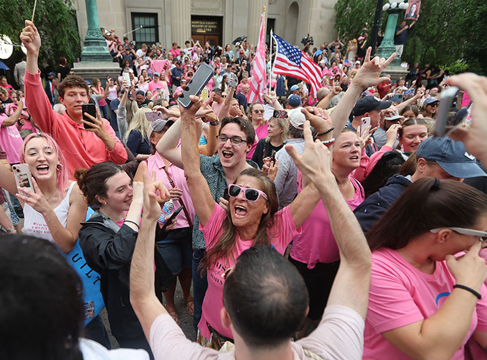 Crowd celebrating Karen Read acquittal in trial for slaying cop boyfriend, many wearing pink and holding phones and flags. Crowd celebrating Karen Read acquittal in trial for slaying cop boyfriend, many wearing pink and holding phones and flags.