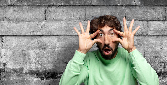 Man in green sweater making wide eye gesture with hands near eyes against a gray wall.